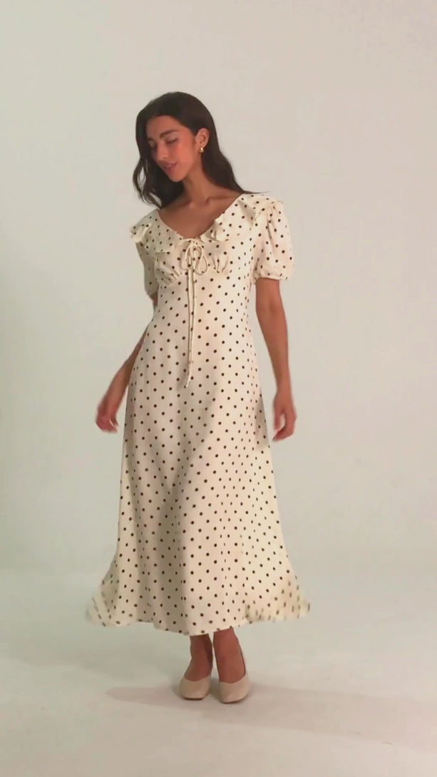 Woman wearing a white midi dress with black polka dots, puff sleeves, and a ruffled neckline with bow tie, facing forward with a slight smile