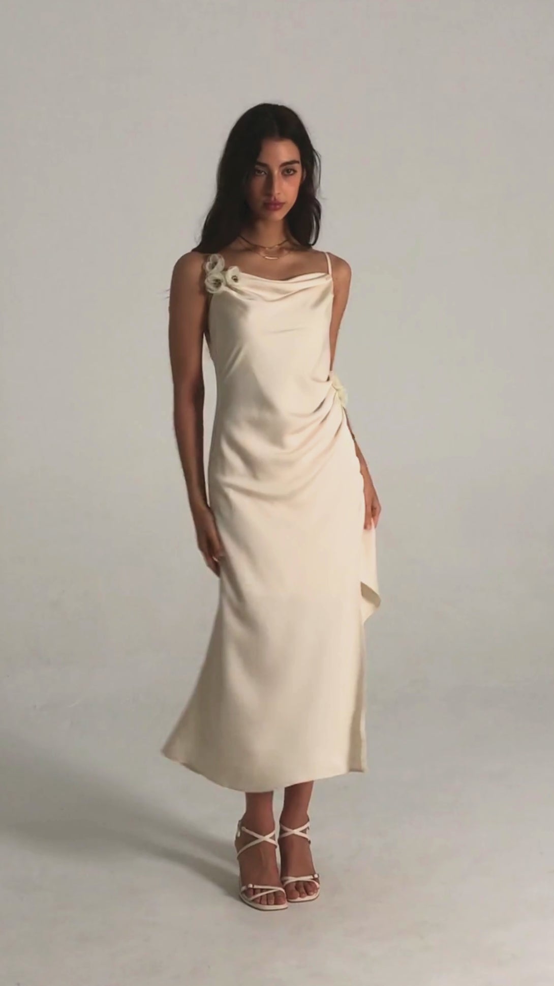 Front view of a white satin midi dress with cowl neckline, delicate floral appliqués at the strap and waist, side slit, and a fluid drape