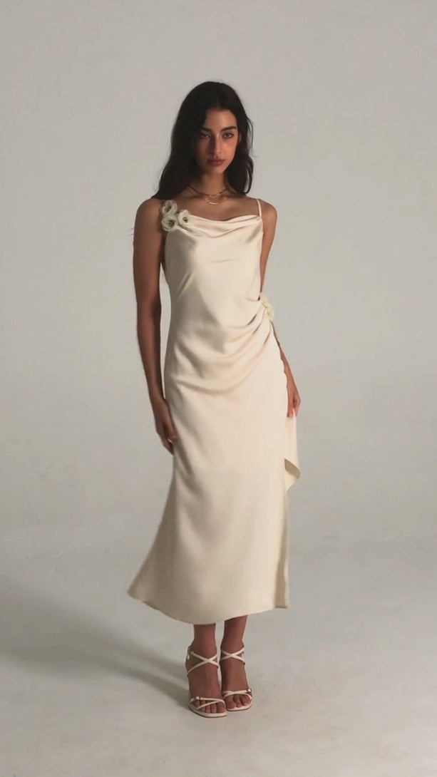 Front view of a white satin midi dress with cowl neckline, delicate floral appliqués at the strap and waist, side slit, and a fluid drape
