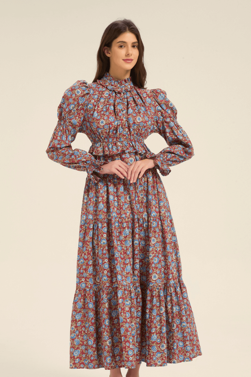Model wearing a red floral long sleeve maxi dress with puffed shoulders and cinched waist, standing with hands clasped.