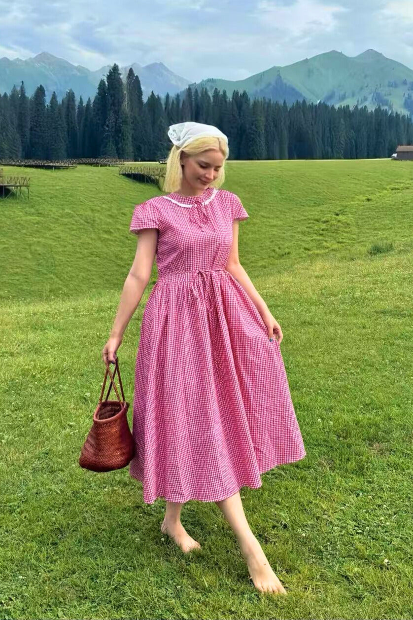 Woman in red gingham midi dress walking barefoot on grass, holding a woven bag, styled with a white headscarf in a countryside setting