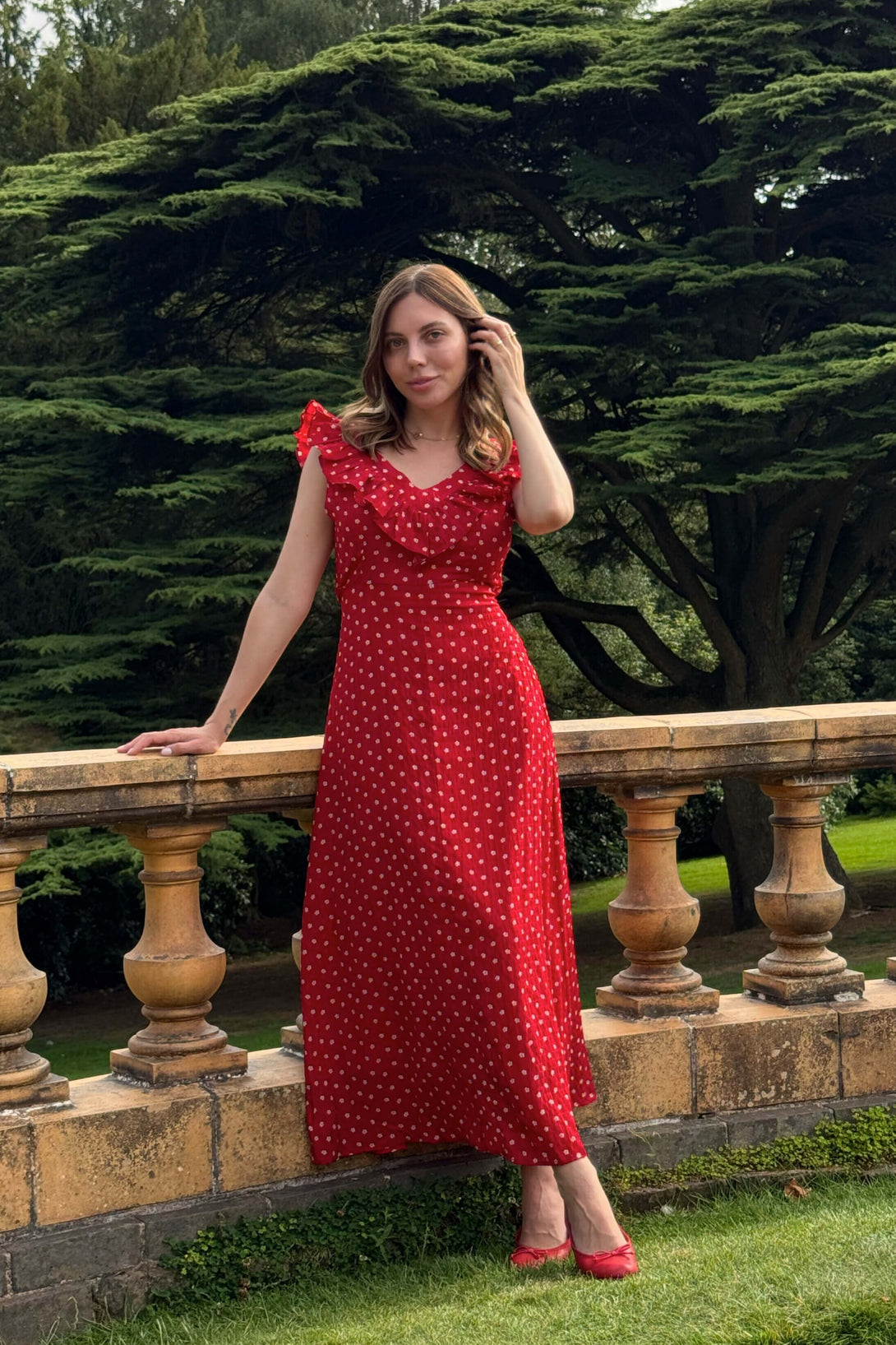 Woman wearing a red polka dot maxi dress with ruffle neckline, posing outdoors by a stone railing with lush green trees in the background.