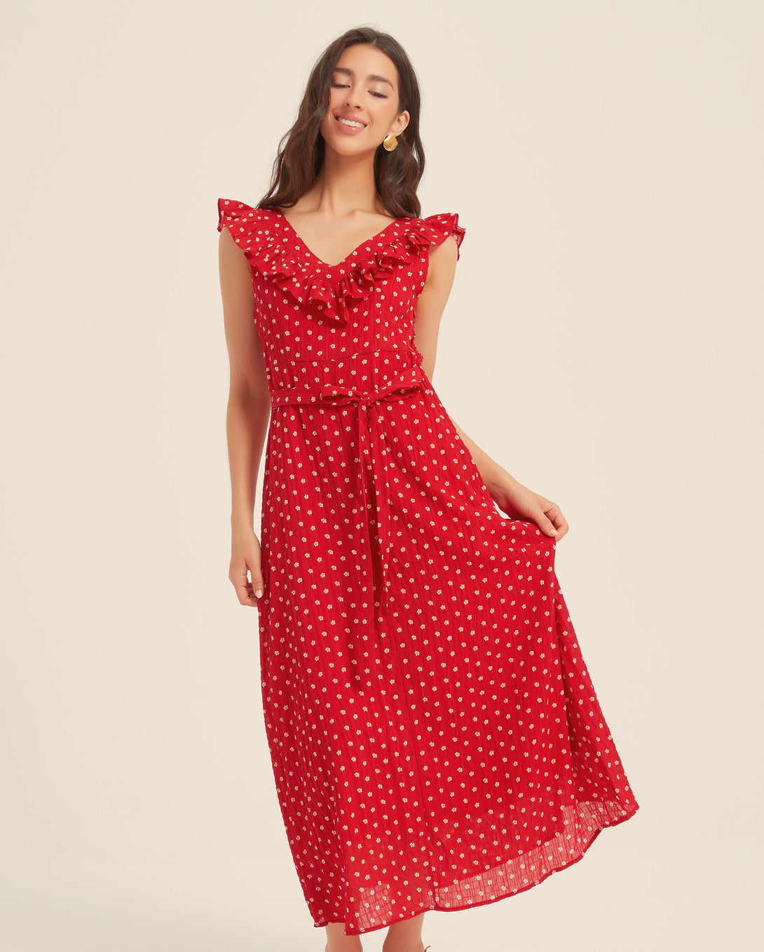 Smiling woman wearing a red ruffle polka dot midi dress with tie waist, holding the skirt hem in a light, joyful pose against a neutral background
