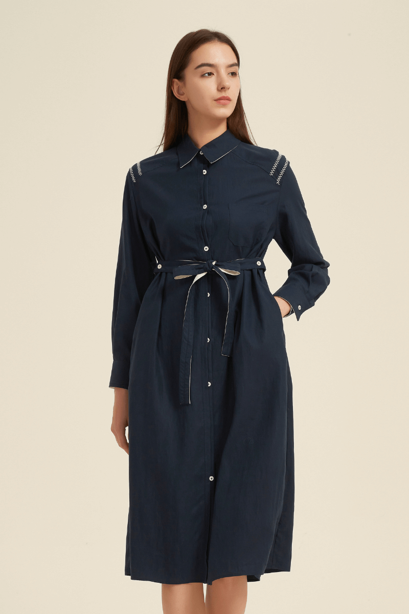 Side front view of a navy midi shirt dress with a collared neckline, button-down closure, belted waist, long sleeves, and contrast stitching at the shoulders.
