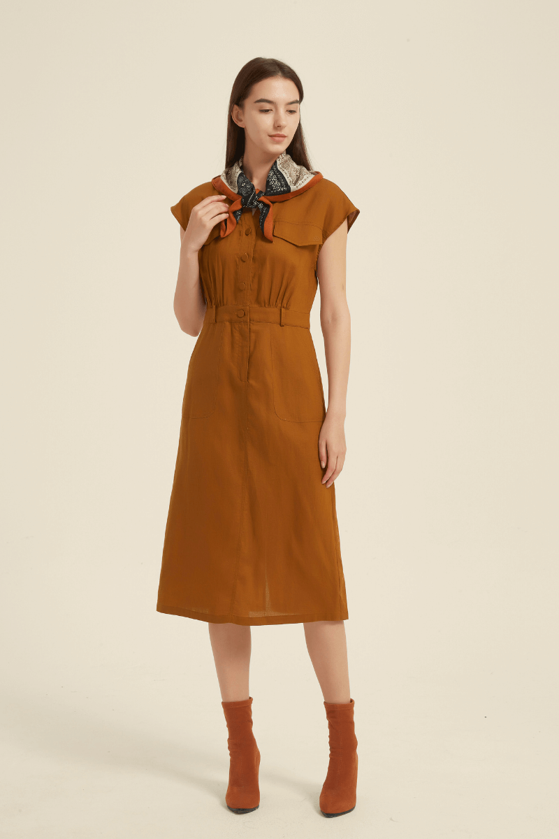 Side front view of a rust cap sleeve midi shirt dress with a button-down front, elastic waist, large chest flap pocket, and a printed scarf tied at the neck.