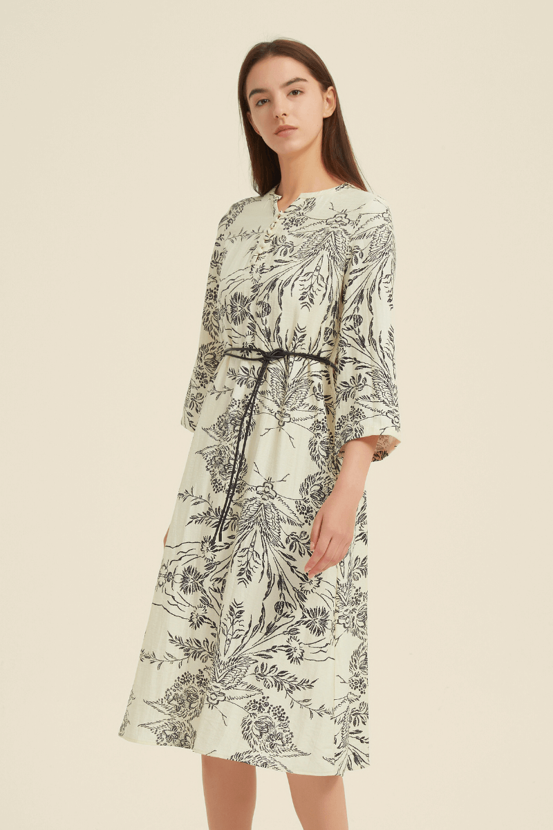Side view of a cream midi dress adorned with a black botanical print, flared sleeves, and a slim black belt for a cinched waist.