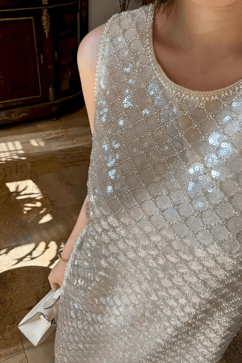 Close-up of a sleeveless silver sequin mini dress featuring pearl-trim neckline and shimmering circular sequin pattern, paired with a white clutch.