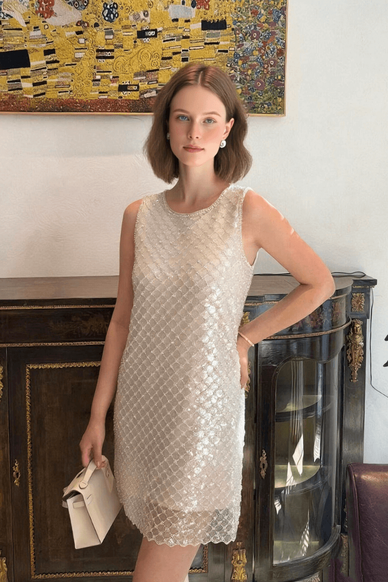 Woman wearing a sleeveless silver sequin mini dress featuring a lattice pattern, sheer scalloped hem, and round neckline, styled with a white clutch in an elegant indoor setting.