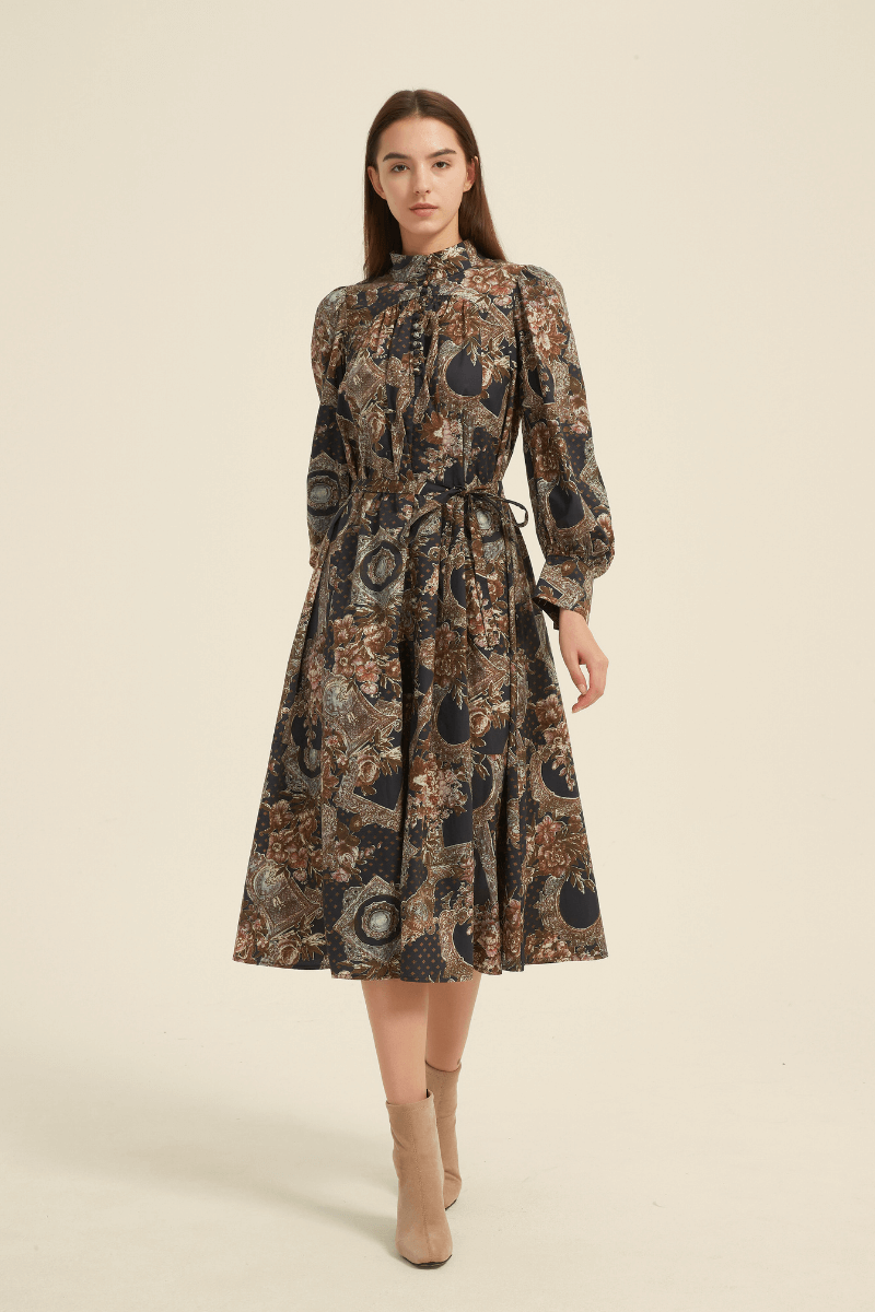Model wearing a vintage-inspired baroque print midi dress in navy, brown, and gold tones, featuring long puff sleeves, a high neckline with fabric-covered buttons, and a self-tie belt, styled with beige ankle boots.