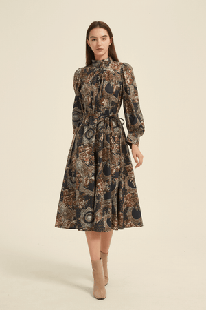 Model wearing a vintage-inspired baroque print midi dress in navy, brown, and gold tones, featuring long puff sleeves, a high neckline with fabric-covered buttons, and a self-tie belt, styled with beige ankle boots.