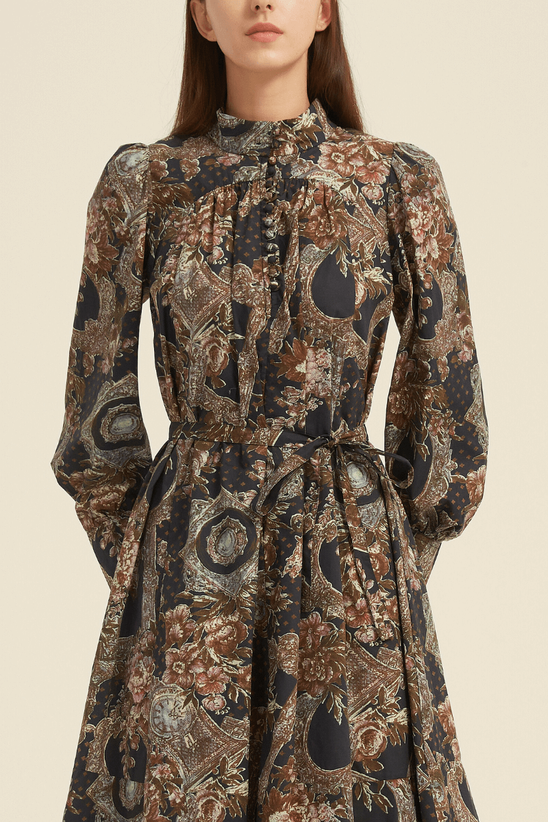 Close-up of a vintage baroque-print midi dress featuring a high ruffled neckline, fabric-covered buttons, and a waist tie, in earthy tones of brown, gold, and navy with ornate floral and frame motifs.