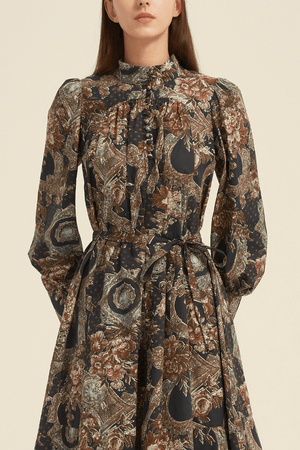 Close-up of a vintage baroque-print midi dress featuring a high ruffled neckline, fabric-covered buttons, and a waist tie, in earthy tones of brown, gold, and navy with ornate floral and frame motifs.