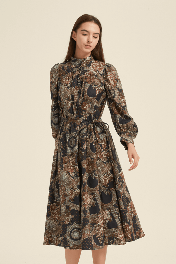 Front view of a vintage baroque-print midi dress featuring long puff sleeves, a high neckline with button details, a waist tie, and an A-line skirt in ornate floral and frame patterns of brown, gold, and navy.