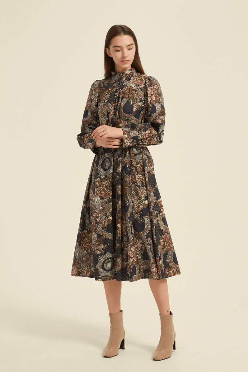 Model wearing a vintage-inspired high-neck midi dress with a baroque floral print in deep earthy tones, long puff sleeves, button-up bodice, and waist tie, paired with beige ankle boots.