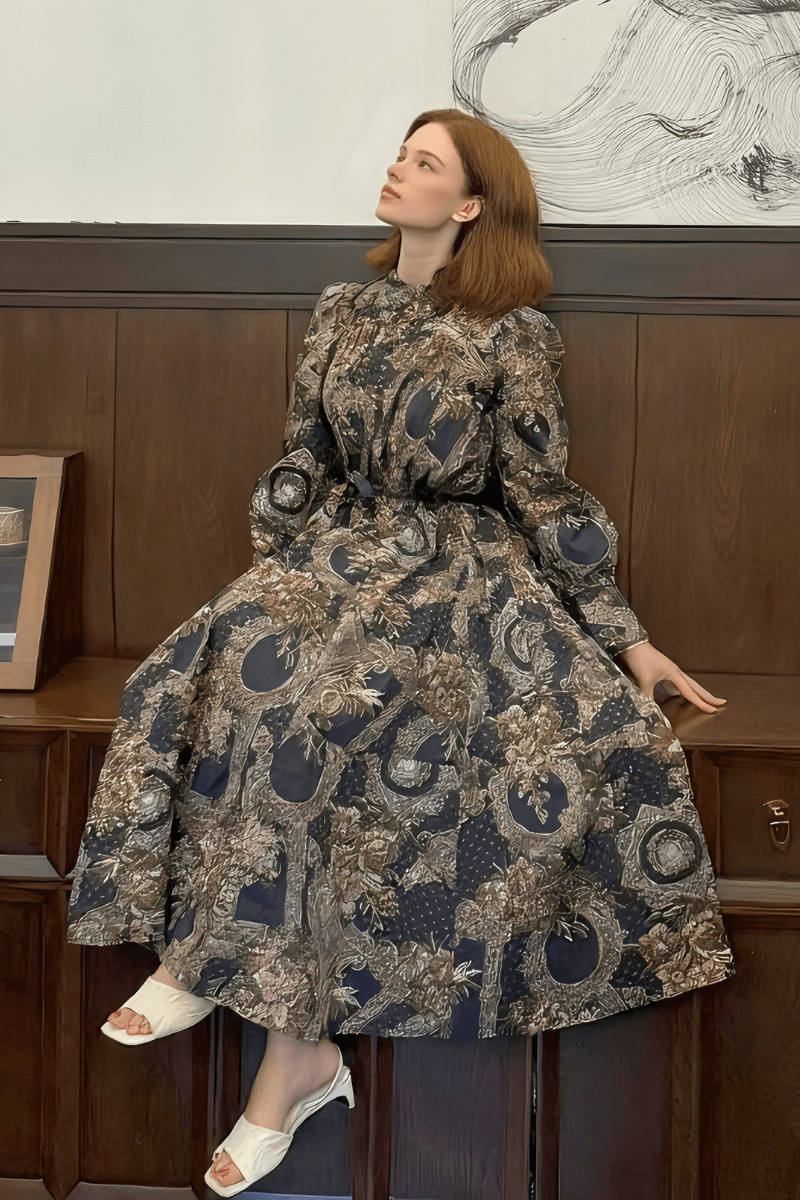 Woman in a vintage print long sleeve maxi dress with a belted waist, featuring earthy brown and navy patterns, sitting elegantly against a wooden wall.