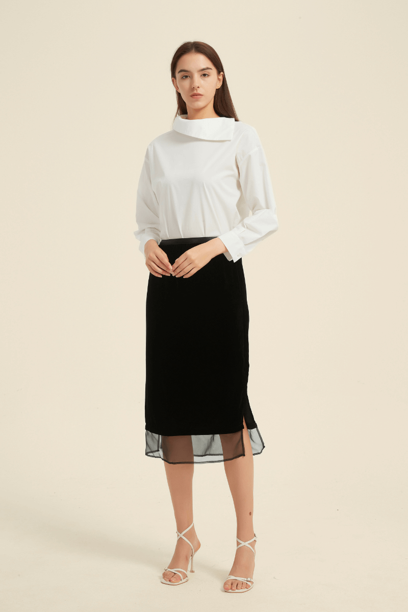 Full front view of a woman wearing a white long-sleeve blouse with an asymmetric collar, paired with a black midi skirt featuring sheer hem panels and side slits.