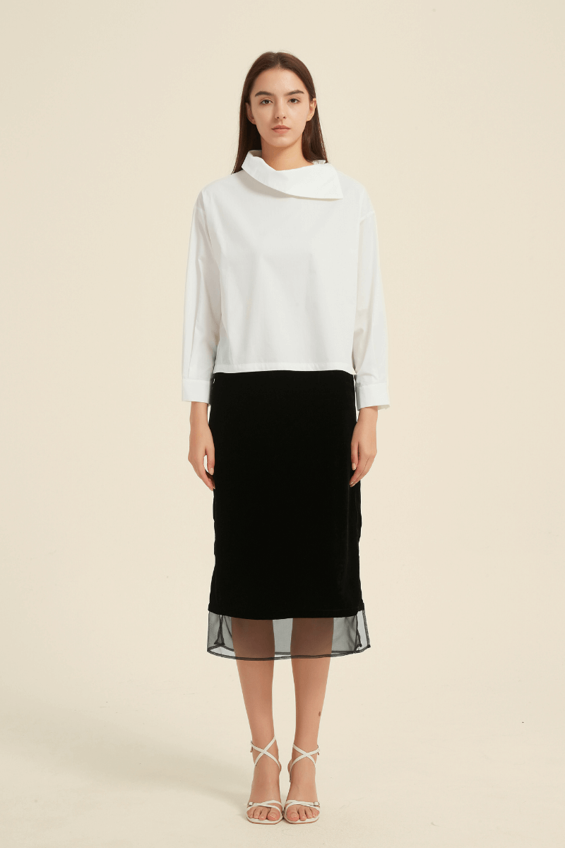 Woman wearing a white long-sleeve blouse with an asymmetric folded collar, paired with a black midi skirt featuring a sheer hemline and strappy heels.