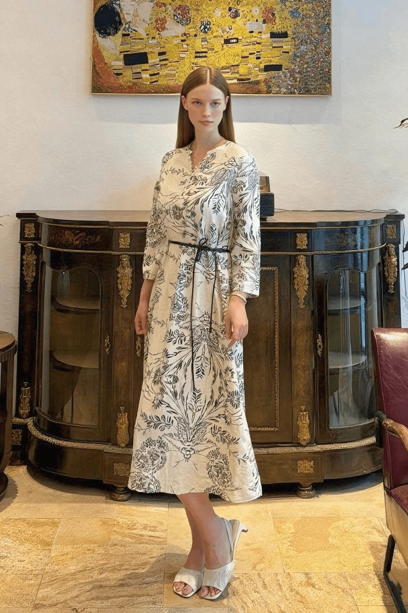 Woman wearing a white midi dress with black floral print, belted waist, and three-quarter sleeves, styled with white heels in an elegant indoor setting.