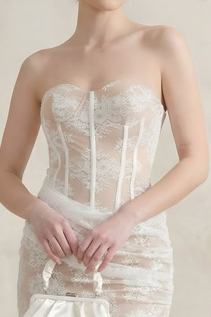 A close-up of a model wearing a white lace strapless corset maxi dress, showing the sheer boned bodice and delicate floral lace texture while she holds a small satin bag.