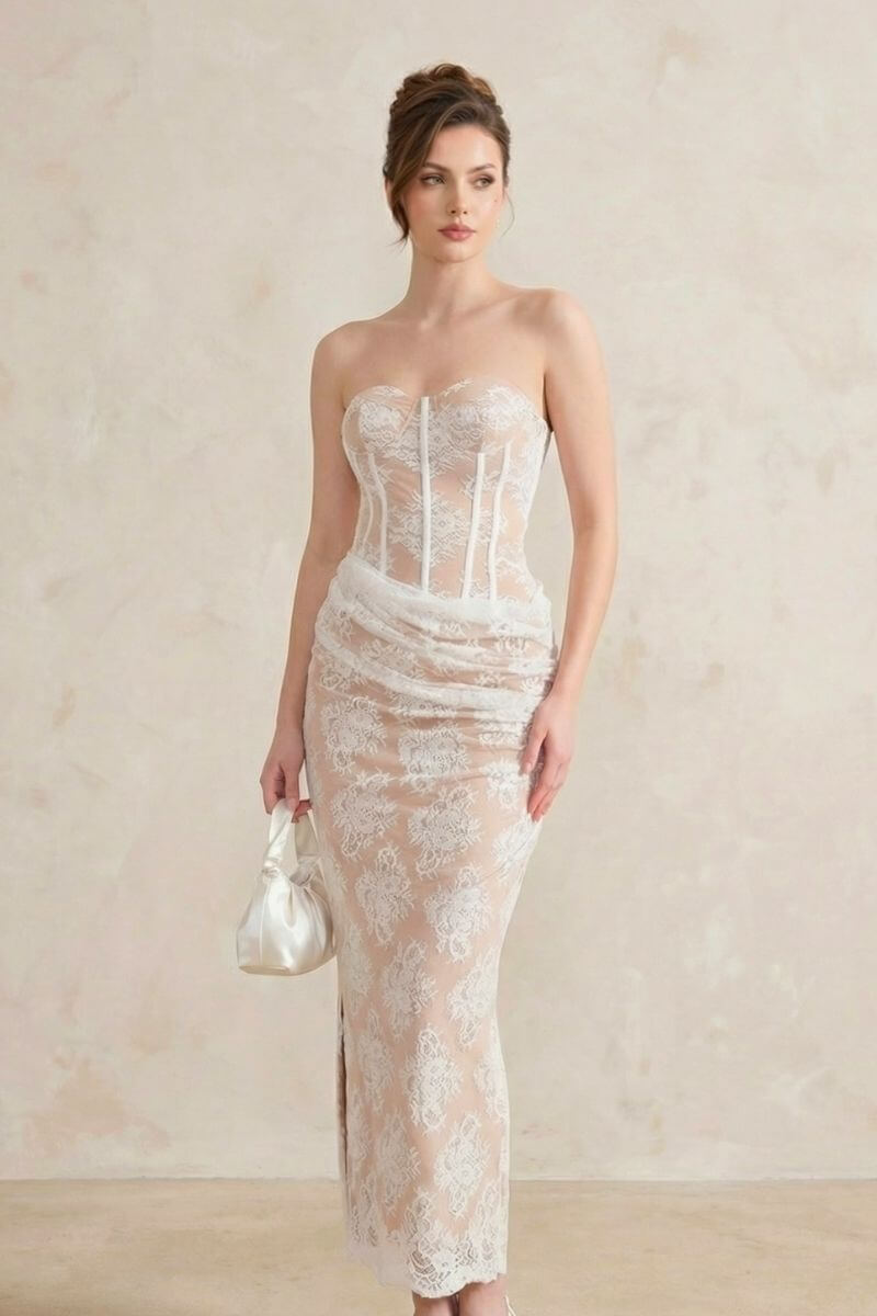 A woman wearing a white lace strapless corset maxi dress holds a satin bag while posing in a minimalist studio.