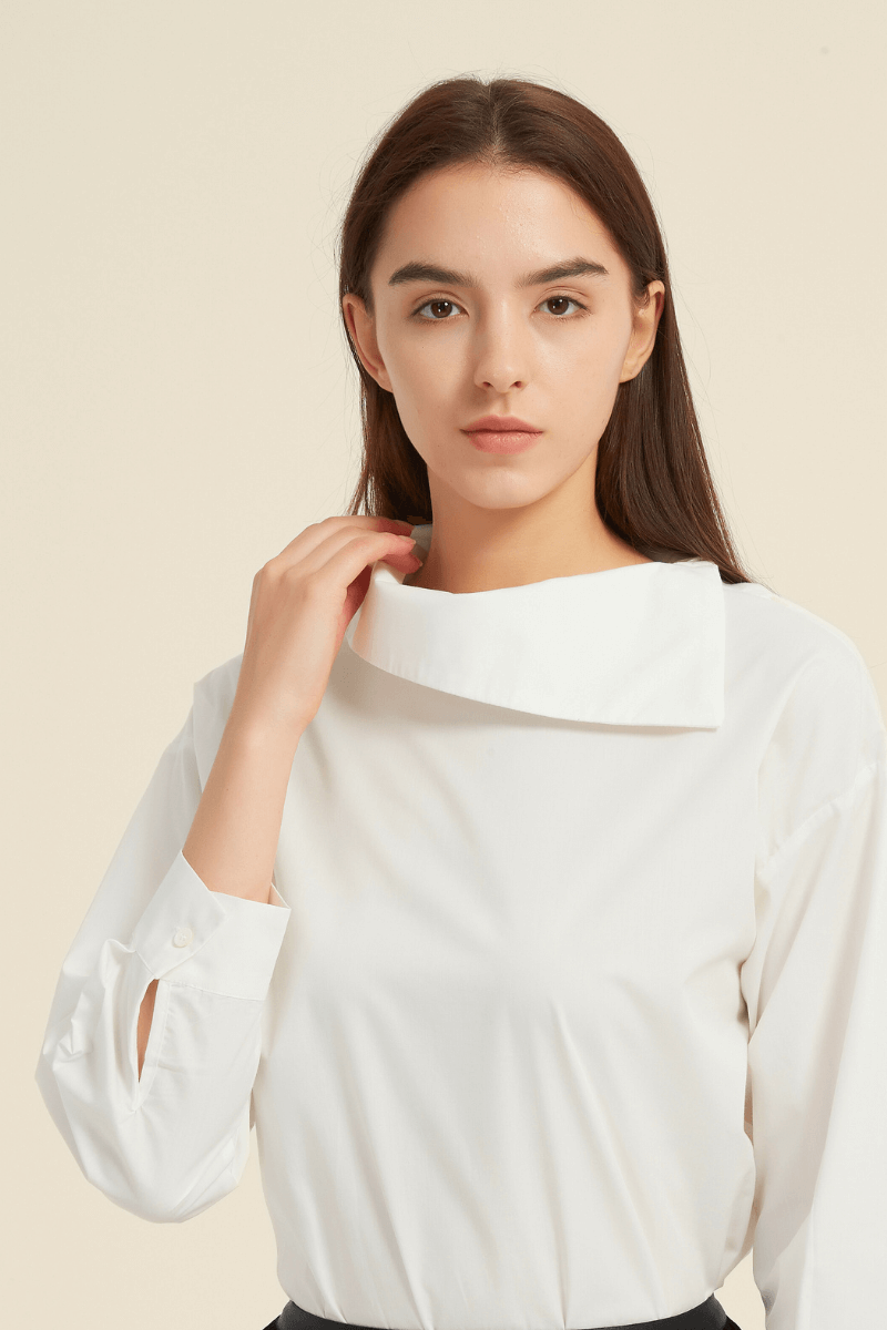 Close-up of a woman wearing a white long-sleeve blouse featuring an asymmetric folded collar and buttoned cuffs, made from smooth lightweight fabric.