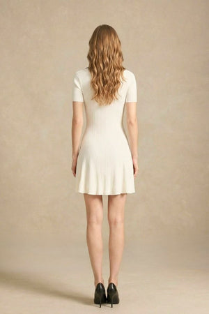 Back view of a white knit mini dress with short sleeves and a flared hem, worn by a woman standing in a neutral studio setting.