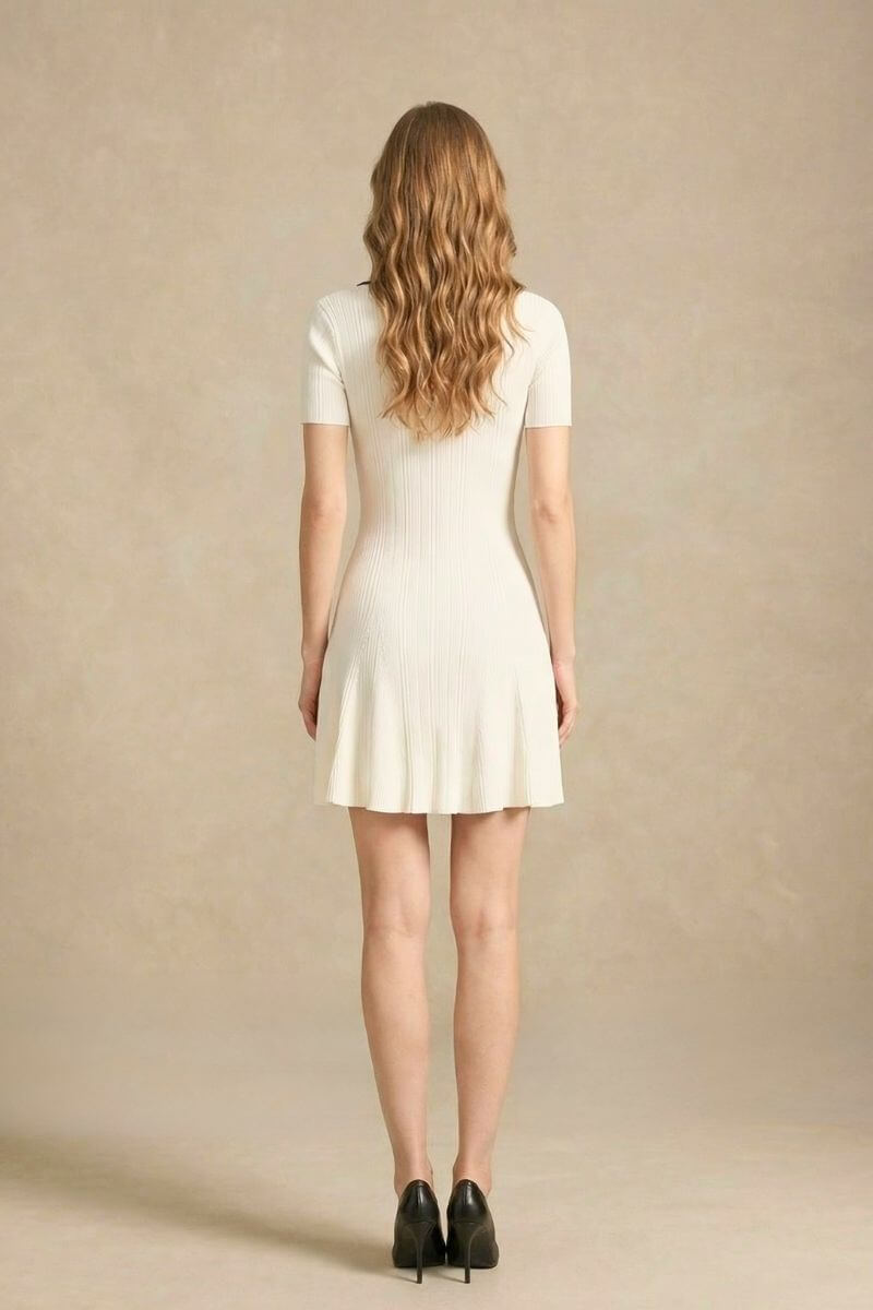 Back view of a white knit mini dress with short sleeves and a flared hem, worn by a woman standing in a neutral studio setting.