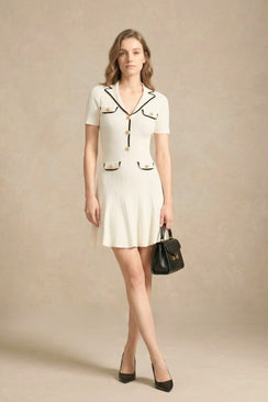 A woman wearing a white knit mini dress with black contrast trim and gold buttons, styled with black heels and a black handbag.