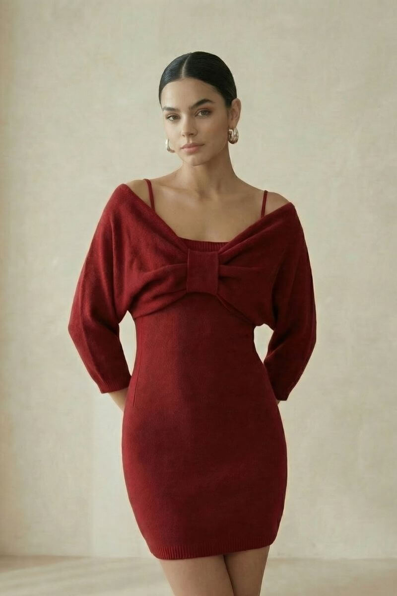 A woman stands in a studio wearing a wine-red knit mini dress featuring an off-shoulder bow design and thin straps, captured in a soft, minimal setting for a refined look.