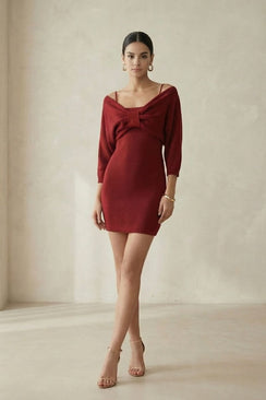 A woman poses in a minimal studio wearing a wine-red off-shoulder knit mini dress with a bow detail and thin straps, highlighting the fitted silhouette and elegant stance.