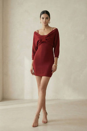 A woman poses in a minimal studio wearing a wine-red off-shoulder knit mini dress with a bow detail and thin straps, highlighting the fitted silhouette and elegant stance.