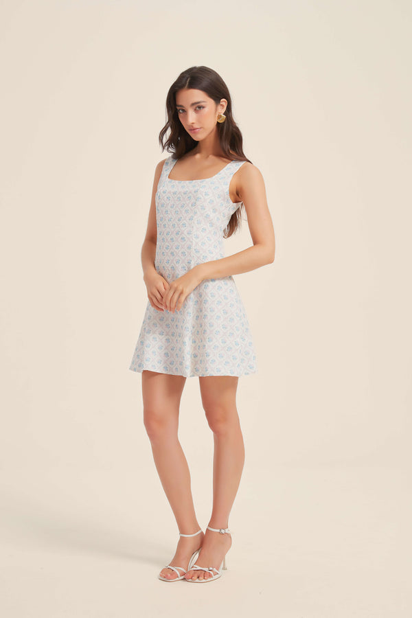 Front view of a woman wearing a sleeveless blue floral mini dress with a square neckline and A-line silhouette, paired with white strappy sandals.