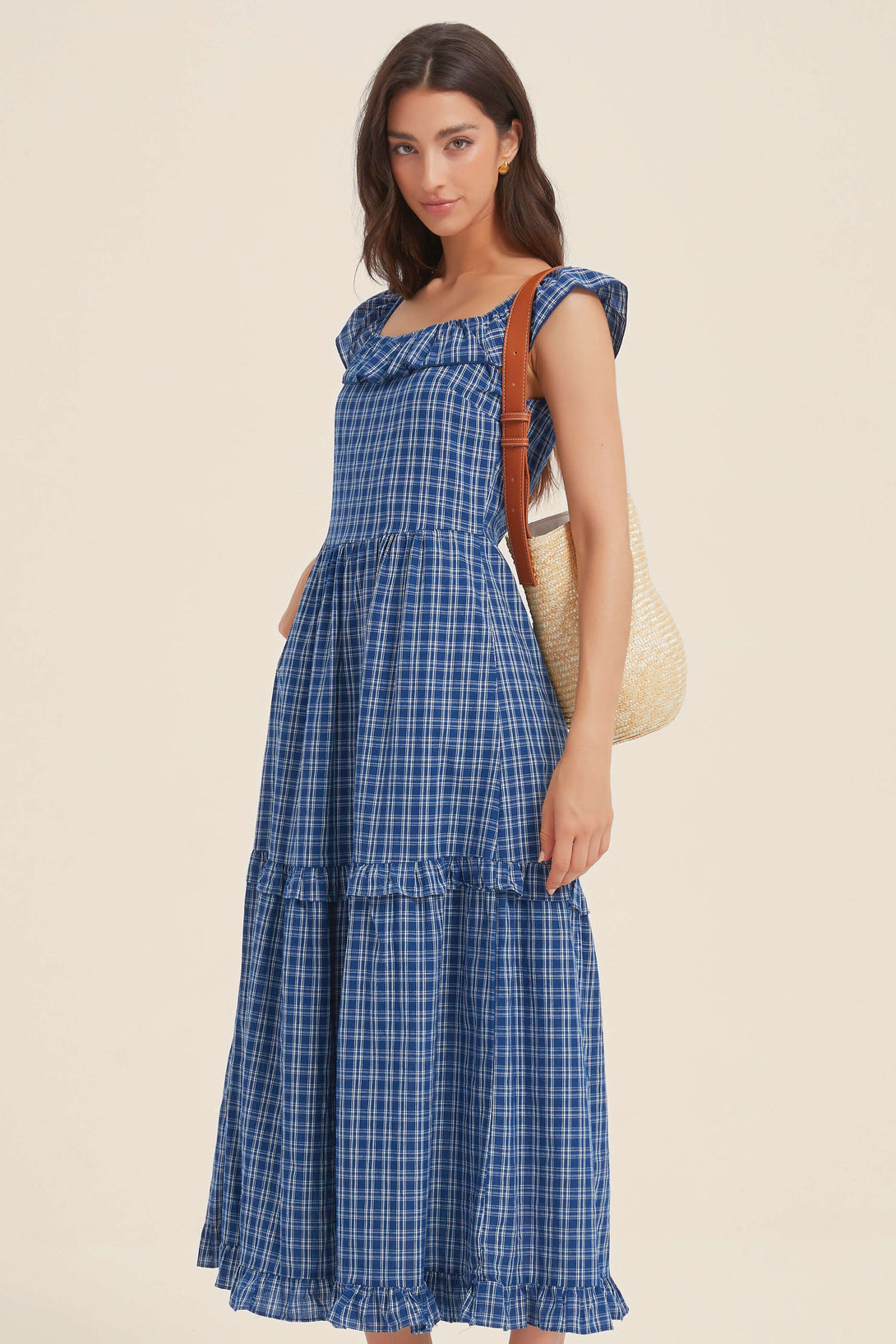 Woman in a blue gingham midi dress with ruffle neckline, styled with a straw shoulder bag for a casual summer outing.