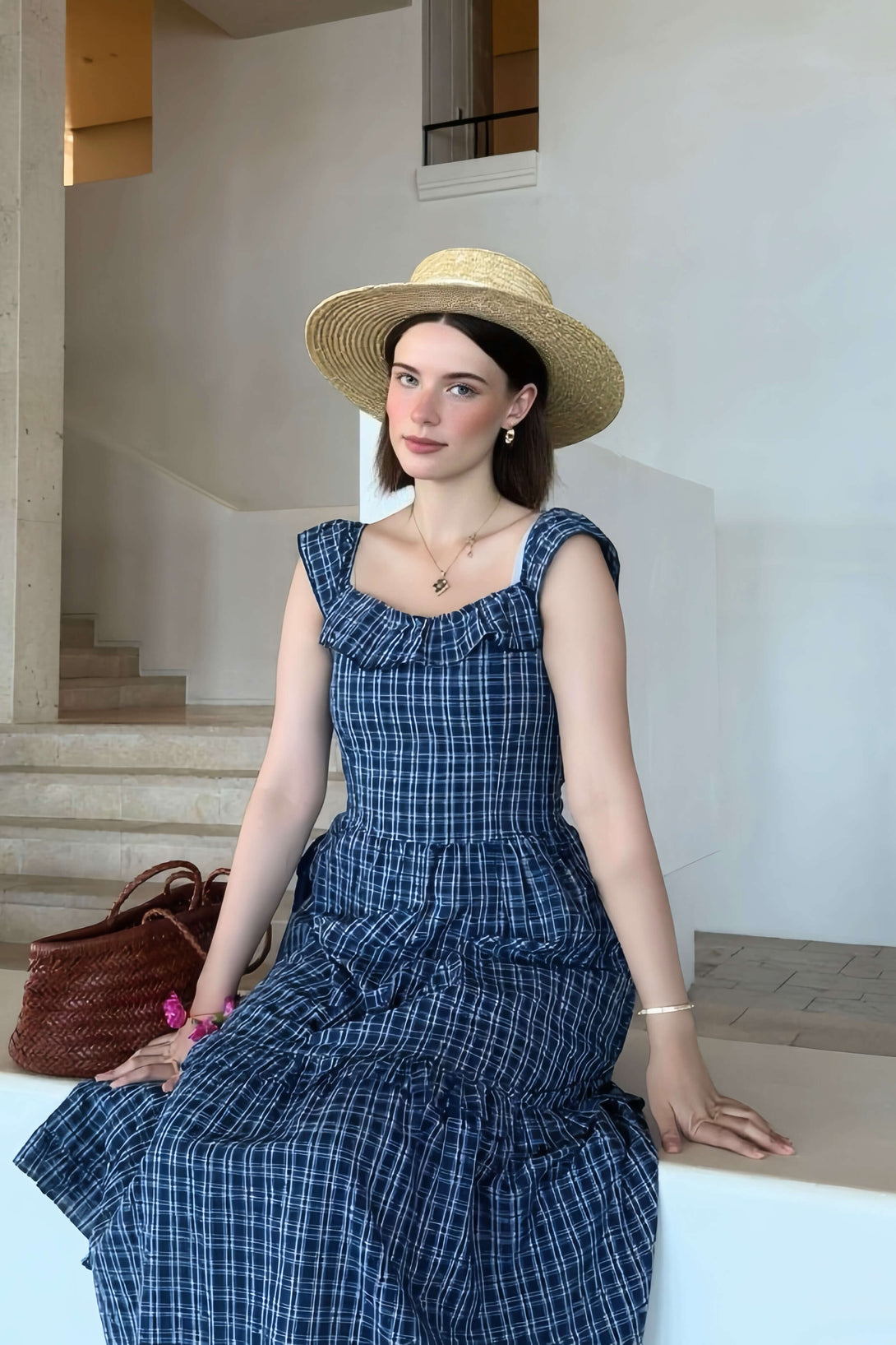 Woman sitting indoors in a blue ruffled gingham midi dress and straw hat, styled with a woven bag for a relaxed summer look.