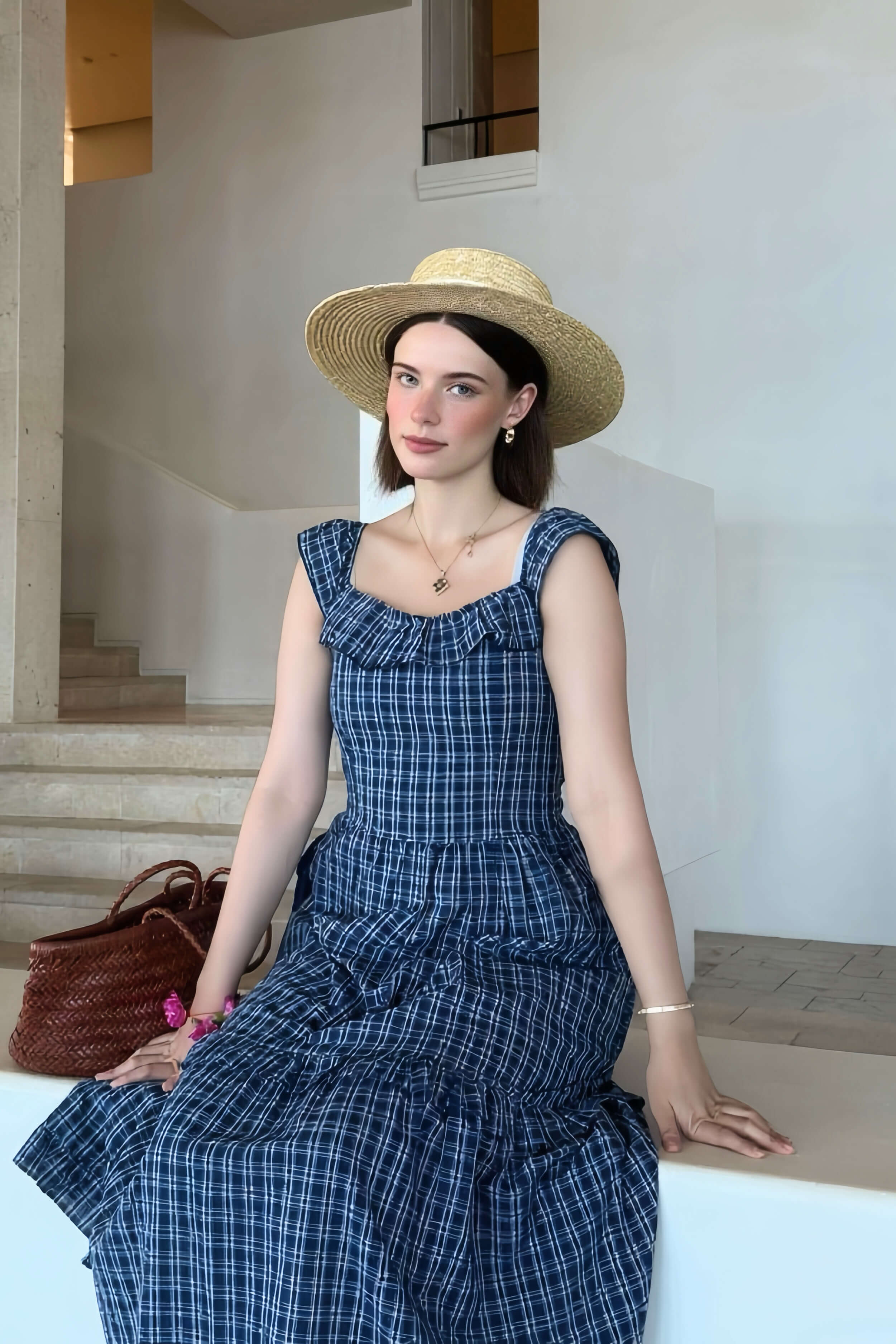 Woman sitting indoors in a blue ruffled gingham midi dress and straw hat, styled with a woven bag for a relaxed summer look.