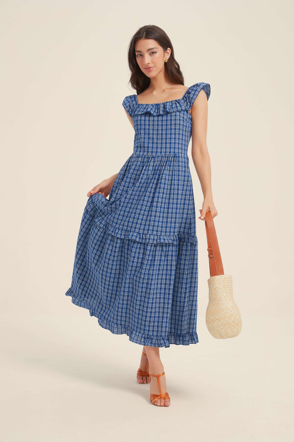 Woman wearing a blue smocked ruffle midi dress with cap sleeves and tiered hem, styled with a straw bag and heels.