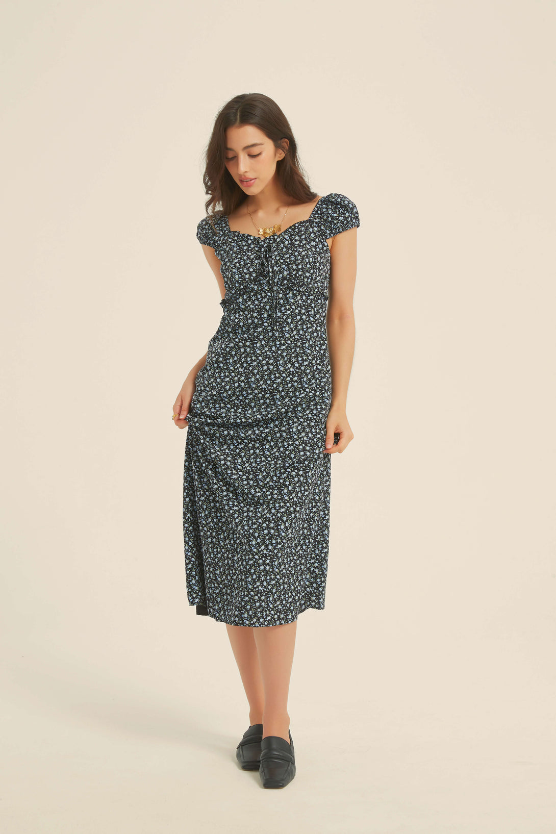 Full front view of a woman wearing a black floral midi dress with cap sleeves and ruched bust, styled with black loafers