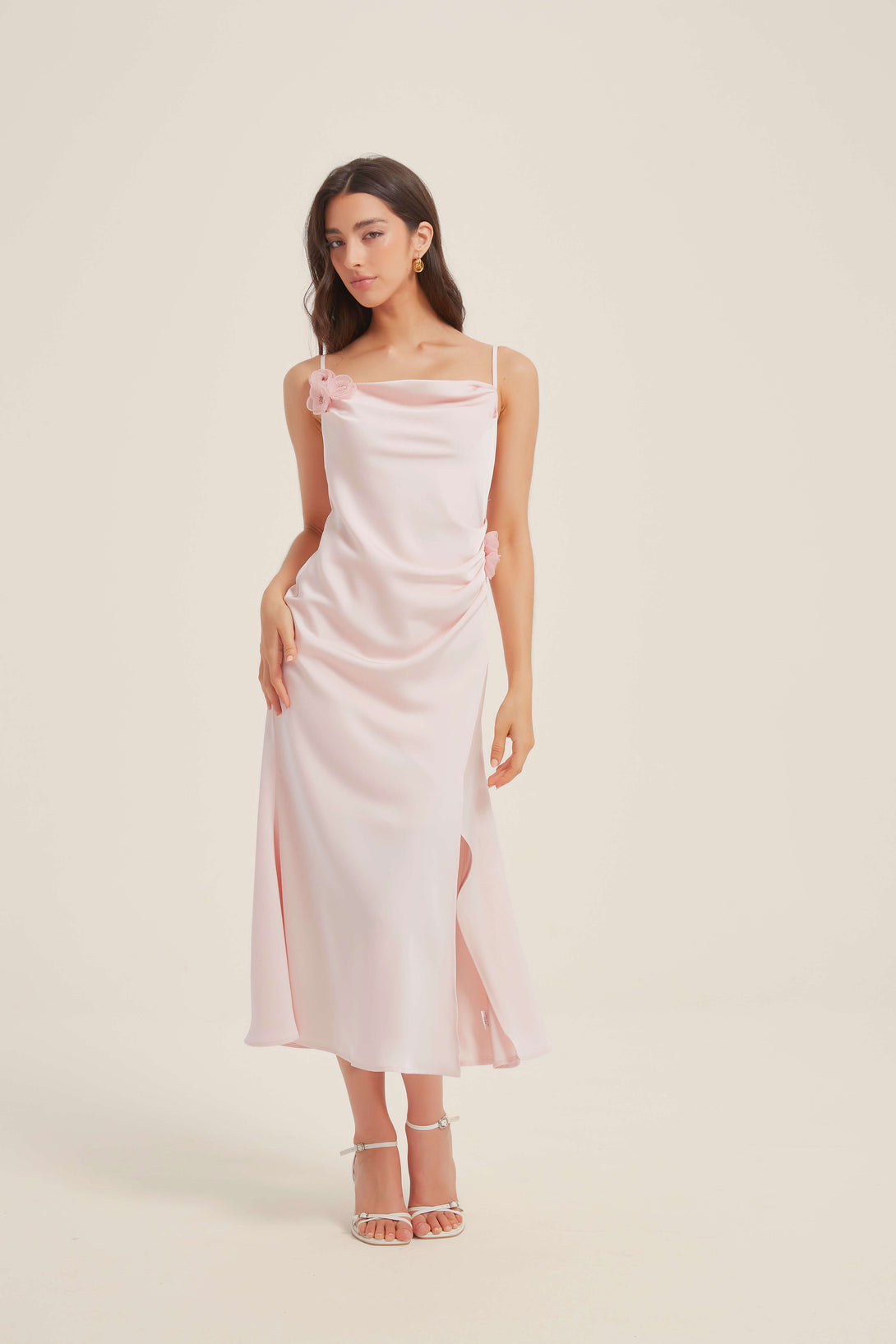 Full front view of a woman wearing a pink satin midi dress with floral appliqué, cowl neckline, and front slit, styled with heels