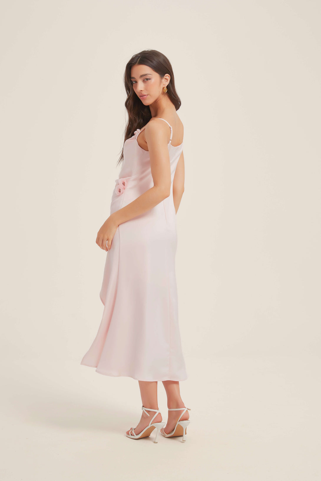 Side view of a pink satin midi dress featuring spaghetti straps, floral rosette detail at the waist, and soft flowing fabric with a subtle flare