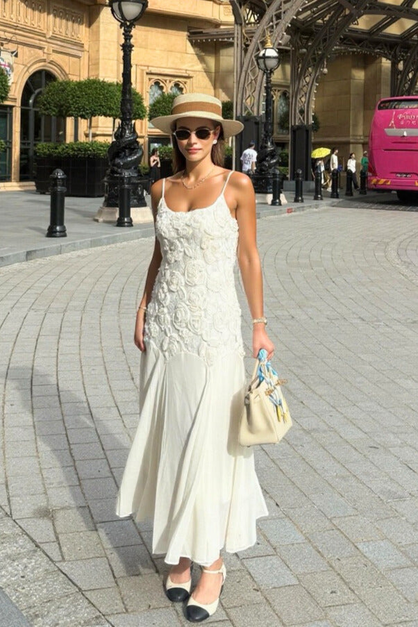 Woman wearing a white floral lace maxi dress styled with a straw hat, sunglasses, and two-tone heels, photographed on a city street.