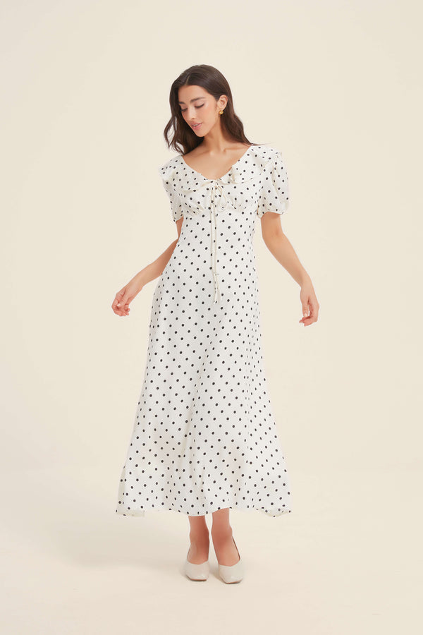 Woman wearing a white polka dot midi dress with puff sleeves and front tie detail, standing gracefully against a light neutral background