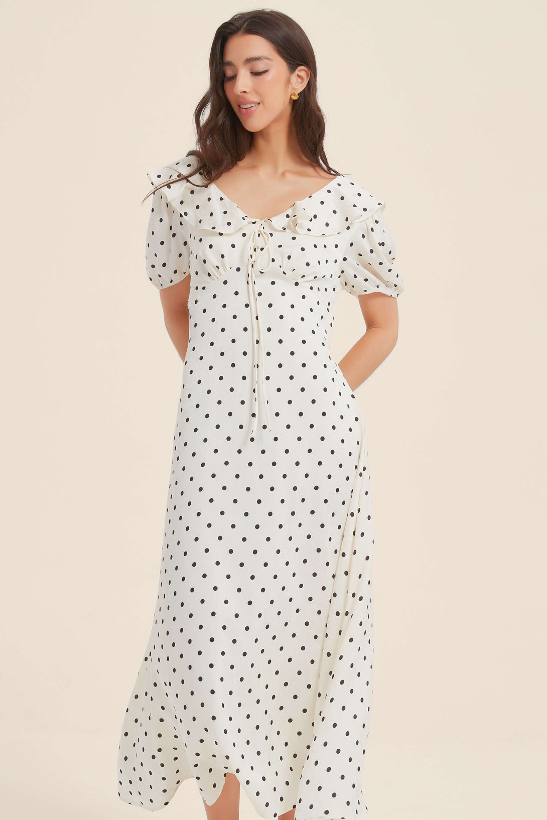 Woman wearing a white midi dress with black polka dots, puff sleeves, and a ruffled neckline with bow tie, facing forward with a slight smile