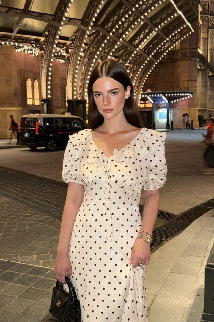 Woman wearing a white polka dot midi dress with puff sleeves and front tie, standing under city lights in a nighttime street scene