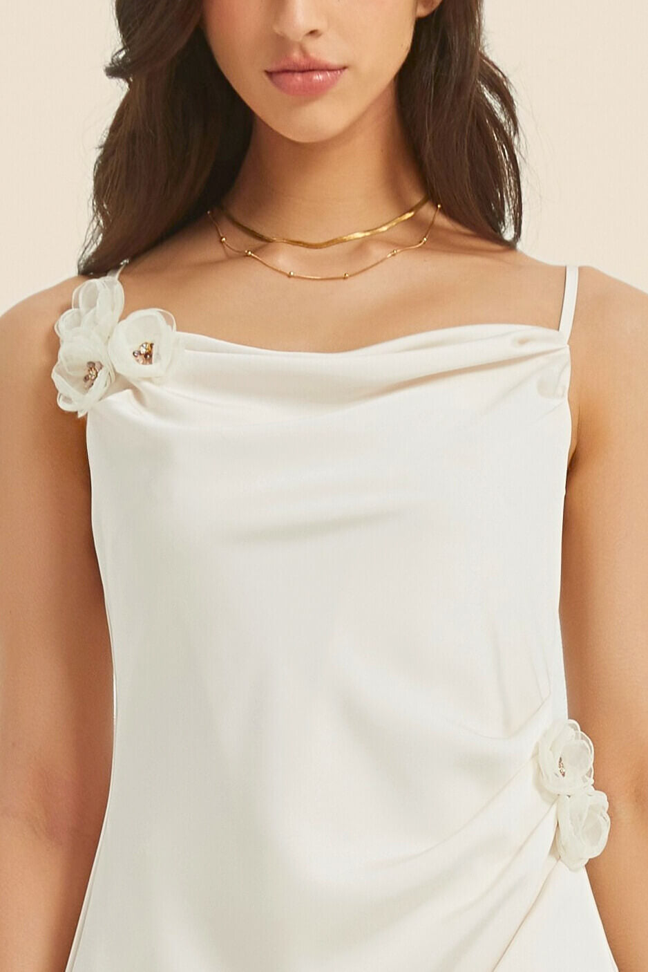 Close-up detail of a white satin midi dress featuring delicate floral appliqués at the strap and waist, with a soft cowl neckline and thin shoulder straps