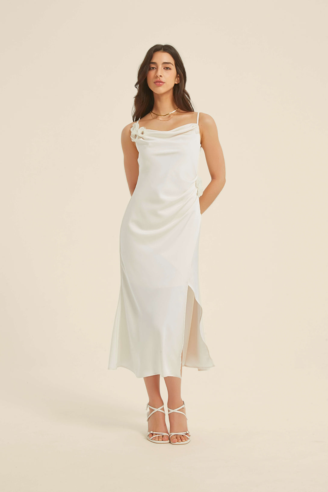 Front view of a white satin midi dress with cowl neckline, delicate floral appliqués at the strap and waist, side slit, and a fluid drape