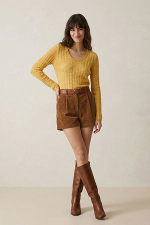 A woman in a yellow ribbed long-sleeve sweater, brown corduroy shorts, and tall brown boots posing full-body in a minimalist studio.